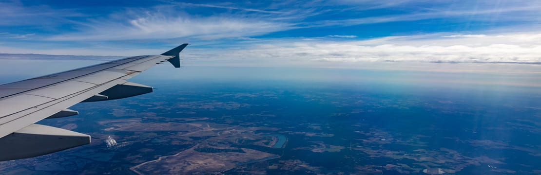 View from airplane window showing wing over a landscape with scattered fields and rivers under a blue sky with wispy clouds.